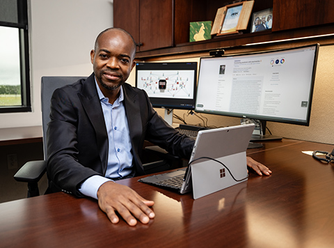 Dr. Martial Ndeffo-Mbah, assistant professor, poses at his desk with research showing on his computer monitors about how smartwatch health sensors can help guard against future pandemics.